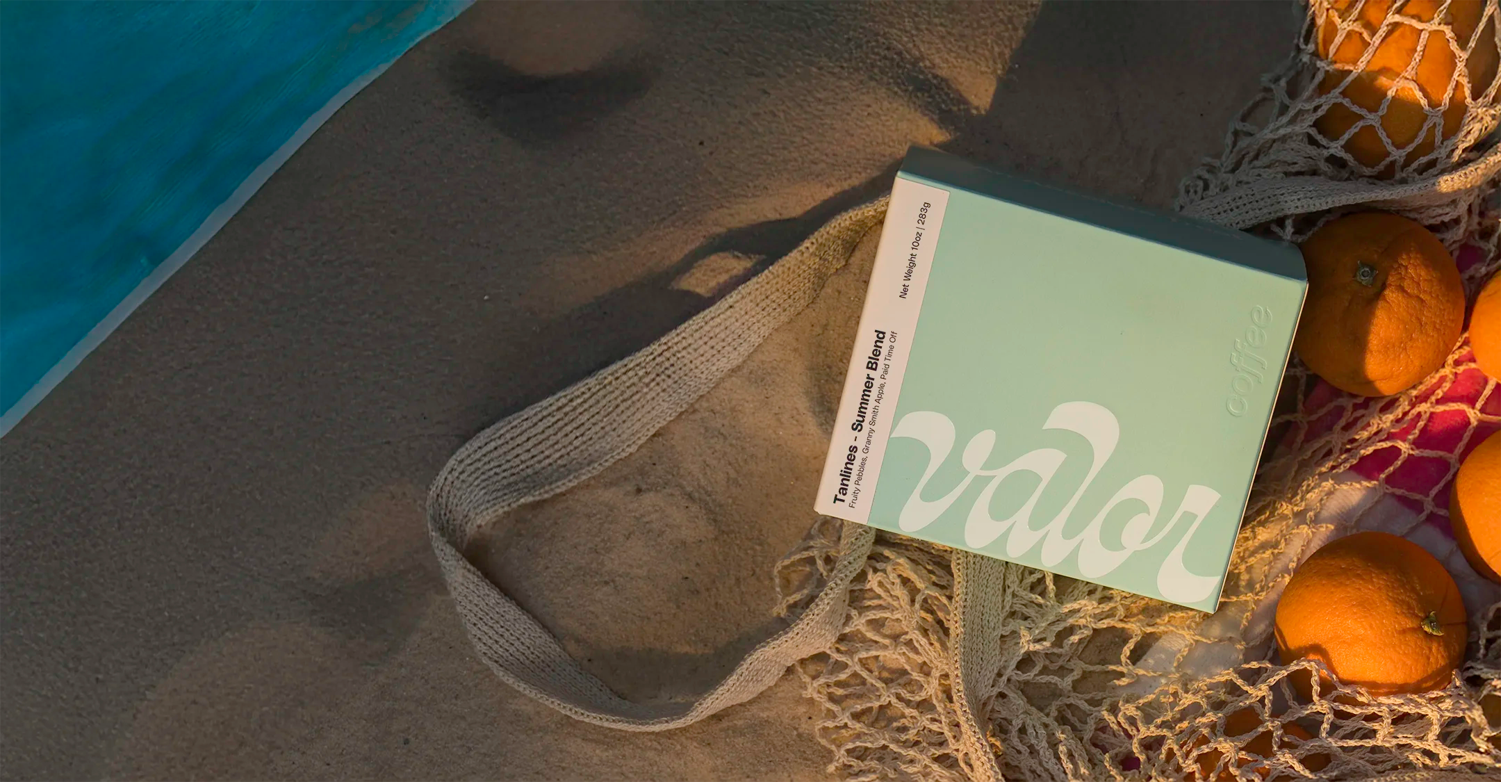 A teal box of Valor's Tanlines Summer Blend sits on a beach under warm, setting sunlight. The box is next to a beach towel and a cotton mesh bag of citrus.